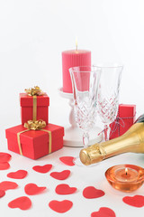 Romantic holiday background: two glasses, a bottle of champagne, red boxes of gifts for your darlings. Decorated with red hearts scattered on a white background and burning candles.