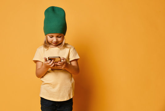 Little Girl Kid In Green Modern Winter Hat Texting In Messenger Using New Popular Mobile Phone On Light Yellow