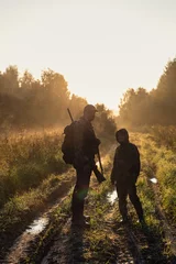 Fototapeten Hunters with hunting equipment going away through rural field towards forest at sunset during hunting season in countryside. © romankosolapov