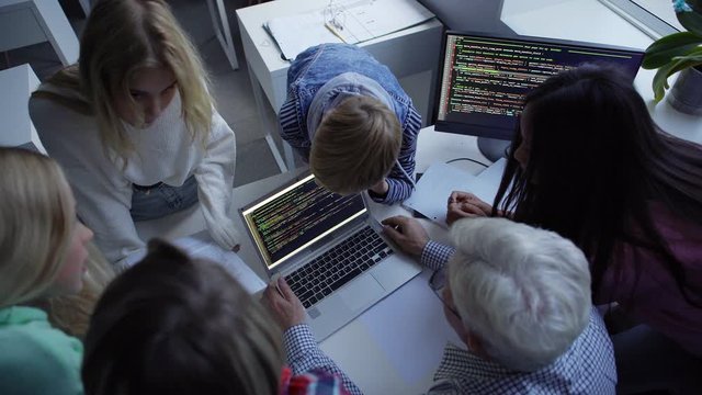 From above of experienced grey haired male teacher sitting at table with laptop and computer and explaining program codes to group of students standing around during computer science lesson
