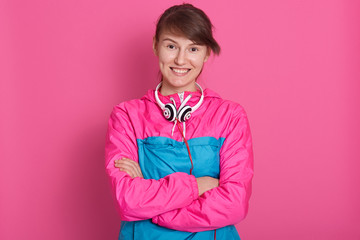 Close up portrait of pretty brunette wears pink sports shirt folded her arms over her chest and looking directly at camera. Lady after fitness, looks happy posing isolated over pink srudio background.