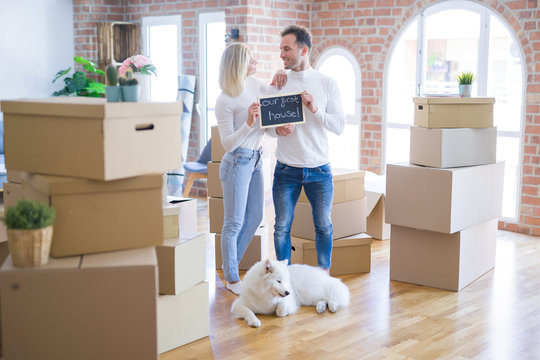 Young beautiful couple with dog standing holding blackboard with message at new home around cardboard boxes
