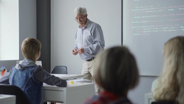 Pan left view of old teacher with pen standing by projector screen and explaining computer science and coding basics to kids in school class
