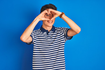 Teenager boy wearing casual t-shirt standing over blue isolated background Doing heart shape with hand and fingers smiling looking through sign