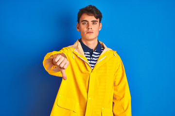 Teenager fisherman boy wearing yellow raincoat over isolated background looking unhappy and angry showing rejection and negative with thumbs down gesture. Bad expression.