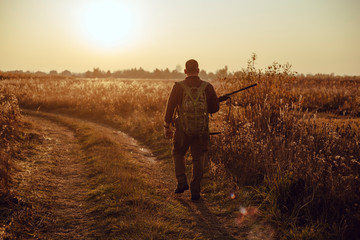 Young hunter in the checkered shirt with military backpack and a gun in his hands walking by the track country road between two fields in the sunset - perfect tiime for duck hunt