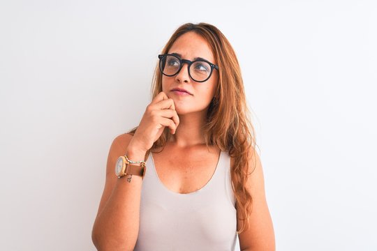Young redhead woman wearing glasses standing over white isolated background with hand on chin thinking about question, pensive expression. Smiling with thoughtful face. Doubt concept.