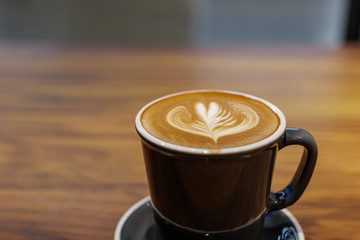 Close up and selected focus view of one cup of cappuccino with small heart and leave pattern of latte art in black ceramic cup on wooden table. Hipster vibes and vintage tone.