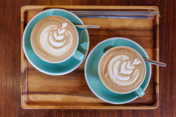 Top flat view of two cups of cappuccino with small heart and leave pattern of latte art in pale green ceramic cups on wooden tray and background.