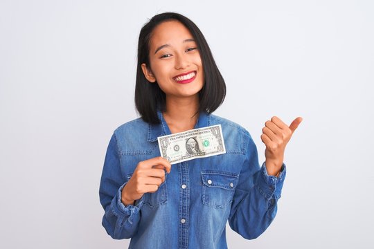 Young beautiful chinese woman holding one dollar standing over isolated white background happy with big smile doing ok sign, thumb up with fingers, excellent sign