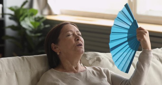 Tired old woman waving fan feel uncomfortable hot at home