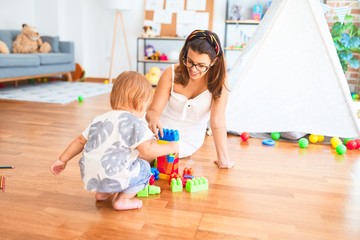 Beautiful teacher and toddler playing with building blocks around lots of toys at kindergarten