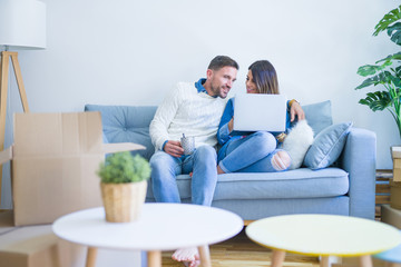 Fototapeta premium Young beautiful couple sitting on the sofa drinking cup of coffee using laptop at new home around cardboard boxes