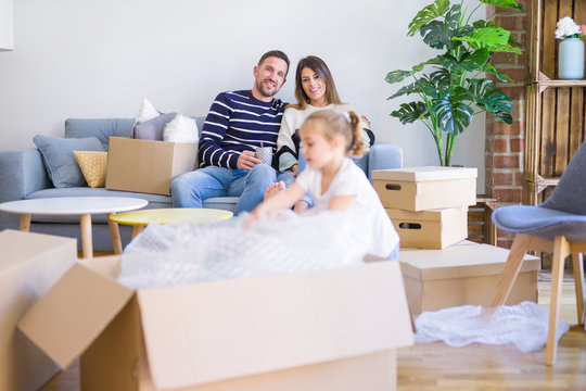 Beautiful family, parents sitting on the sofa drinking coffee looking his kid playing at new home around cardboard boxes