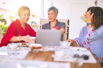 Meeting of middle age women having lunch and drinking coffee. Mature friends smiling happy using laptop at home on a sunny day