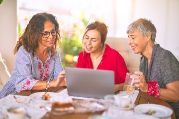 Meeting of middle age women having lunch and drinking coffee. Mature friends smiling happy using laptop at home on a sunny day