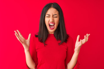 Young beautiful chinese woman wearing casual dress standing over isolated red background celebrating mad and crazy for success with arms raised and closed eyes screaming excited. Winner concept