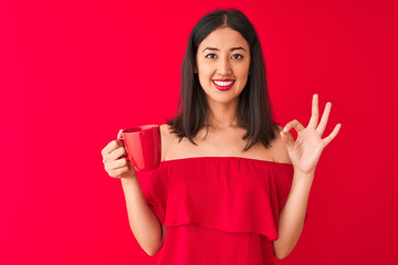 Young beautiful chinese woman drinking cup of coffee standing over isolated red background doing ok sign with fingers, excellent symbol