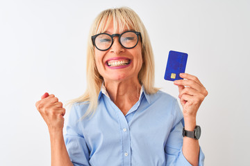Middle age businesswoman wearing glasses holding credit card over isolated white background screaming proud and celebrating victory and success very excited, cheering emotion