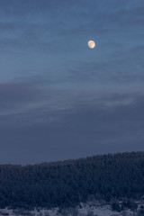 Full moon in the sky with clouds over the evening, winter forest. Dark sky with clouds, a round moon and coniferous forest on top of a snowy mountain. Forest silhouette in hoarfrost.