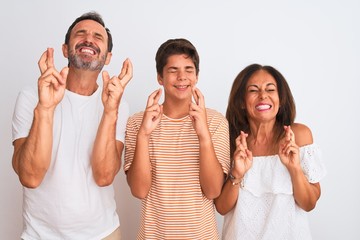 Family of three, mother, father and son standing over white isolated background gesturing finger crossed smiling with hope and eyes closed. Luck and superstitious concept.