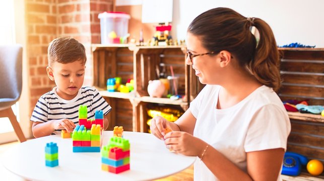 Beautiful teacher and toddler boy playing with construction blocks bulding tower at kindergarten