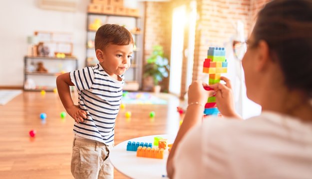 Beautiful teacher and toddler boy playing with construction blocks bulding tower at kindergarten