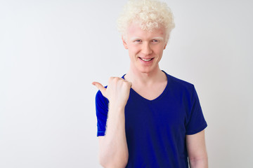 Young albino blond man wearing blue casual t-shirt standing over isolated white background smiling with happy face looking and pointing to the side with thumb up.