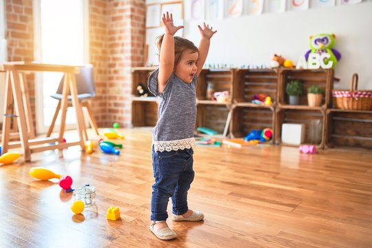 Beautiful toddler standing with hands raised smiling around lots of toys at kindergarten