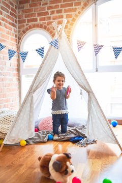 Beautiful toddler standing inside tipi holding plastic dishes smiling at kindergarten