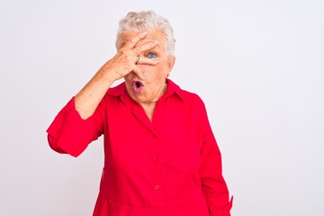 Senior grey-haired woman wearing red casual shirt standing over isolated white background peeking in shock covering face and eyes with hand, looking through fingers with embarrassed expression.