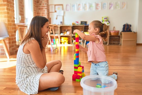 Beautiful teacher and blond toddler girl building tower using plastic blocks at kindergarten