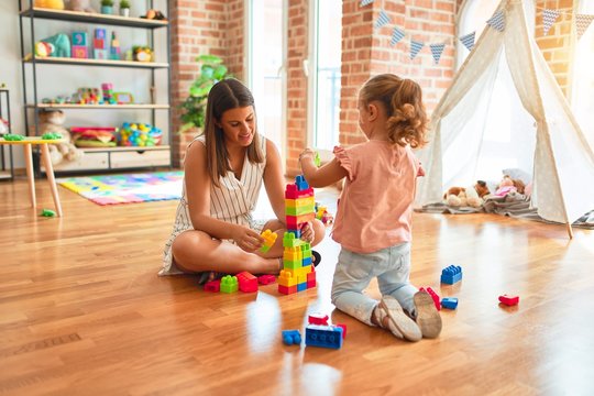 Beautiful teacher and blond toddler girl building tower using plastic blocks at kindergarten
