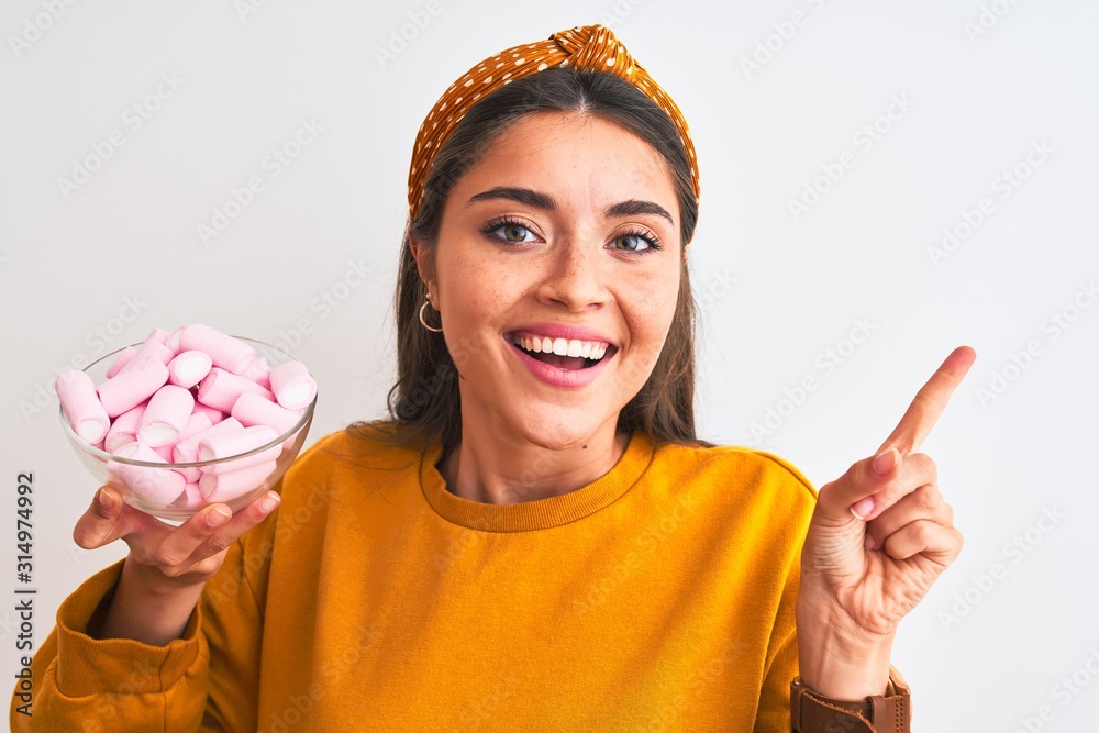 Wall mural Young beautiful woman holding bowl with marshmallows over isolated white background very happy pointing with hand and finger to the side - Wall murals
