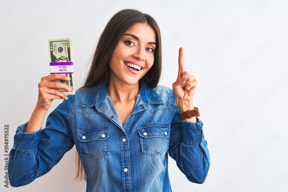 Wall mural Young beautiful woman holding dollars standing over isolated white background very happy pointing with hand and finger to the side - Wall murals