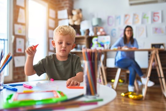 Young caucasian child playing at playschool with teacher. Mother and son at playroom drawing a draw with color pencils, young woman at the background sitting on desk.