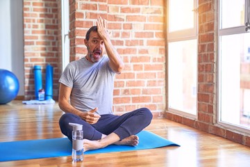 Middle age handsome sportman sitting on mat doing stretching yoga exercise at gym surprised with...