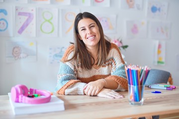Young beautiful teacher woman wearing sweater and glasses sitting on desk at kindergarten with a...