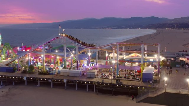Santa Monica Pier, Sep 2019. 4K. Camera follows the rollercoaster train running around the perimeter of Santa Monica amusement park. The variety of rides illuminated with multi-colored lights.