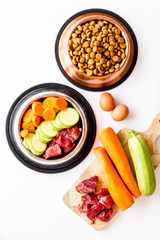Pet feed ingredients. Raw meat and fresh vegetables near bowl with dry feed on white background top-down