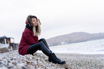 young beautiful caucasian girl in a red warm jacket with a hood, scarf, leather tight pants sitting on a rocky beach enjoying the ocean view