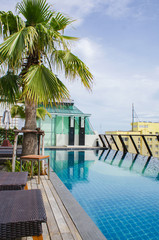 Rooftop pool with sunbeds, a palm tree and showers on a background of blue sky with clouds and a yellow building.