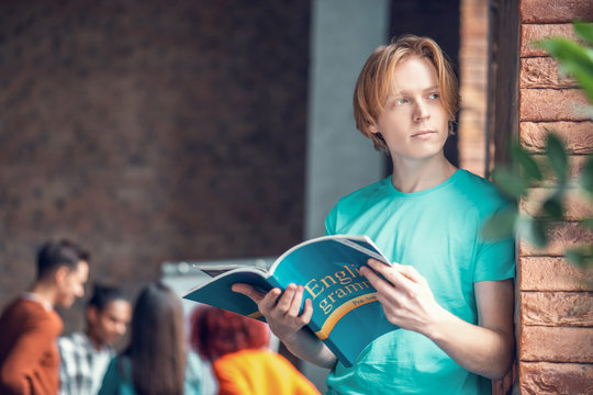 Student standing near brick wall and reading English grammar book