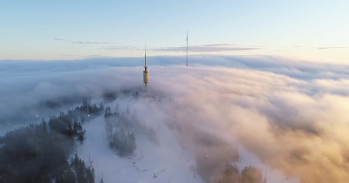 Majestic 4K drone shot with parallax motion of Oslo winter ski park and broadcast tower on top of mountain, with fast moving clouds fog in orange golden sunset, in Tryvann Holmenkollen Oslo Norway.