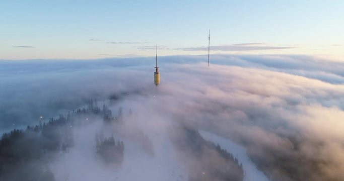 Majestic 4K drone shot with parallax motion of Oslo winter sport park and telecom tower on top of mountain, with fast moving clouds fog in orange golden sunset, in Tryvann Holmenkollen Oslo Norway.