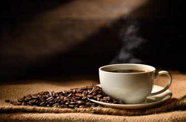 Cup of coffee with smoke and coffee beans on burlap sack on old wooden background