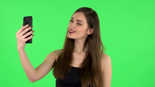 Young woman makes selfie on mobile phone then looking photos on green screen