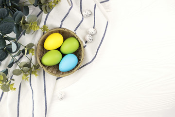 Pastel colored eggs in the nest with the branch of pink flowers on white wooden background. Easter holiday.