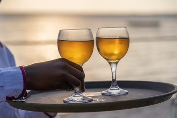 The waiter serves two glasses of white wine on a tray at the beach restaurant during sunset, closeup
