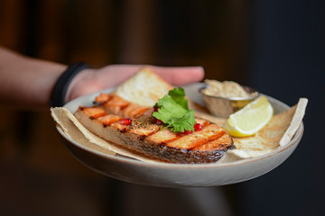 Waiter holds a plate of tasty food, Grilled salmon steak with lemon and pomegranate seeds on a white plate.